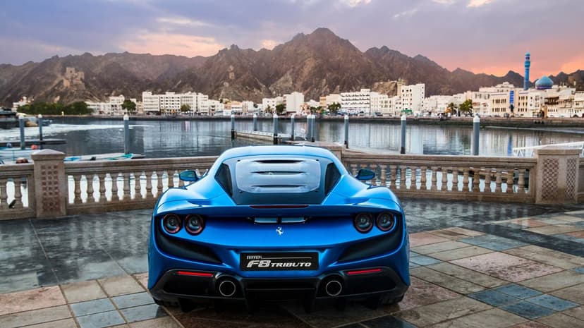 Rear view of a blue Ferrari F8 Tributo on a promenade with mountains and a coastal city at sunset.