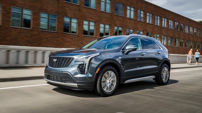 Dark grey Cadillac SUV in motion on a city street with a red brick building and pedestrians in the background.