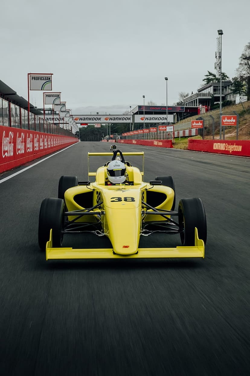 Yellow Formula 1 car numbered 38 navigates a winding asphalt track past red barriers under an overcast sky.