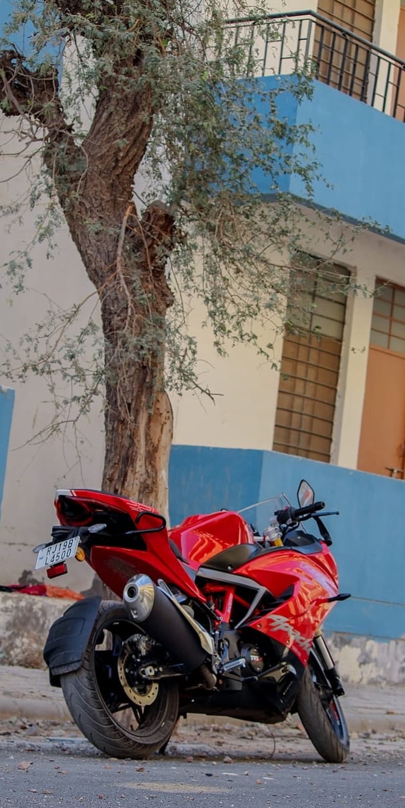 Red sport motorcycle parked on a paved street beside a bare tree and a blue building with barred windows.
