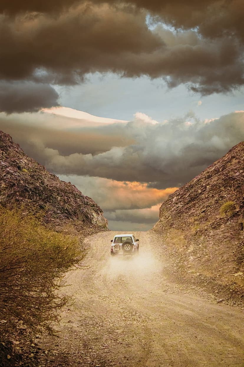 White SUV kicks up dust on a narrow canyon road beneath dark storm clouds and a golden sunset horizon.