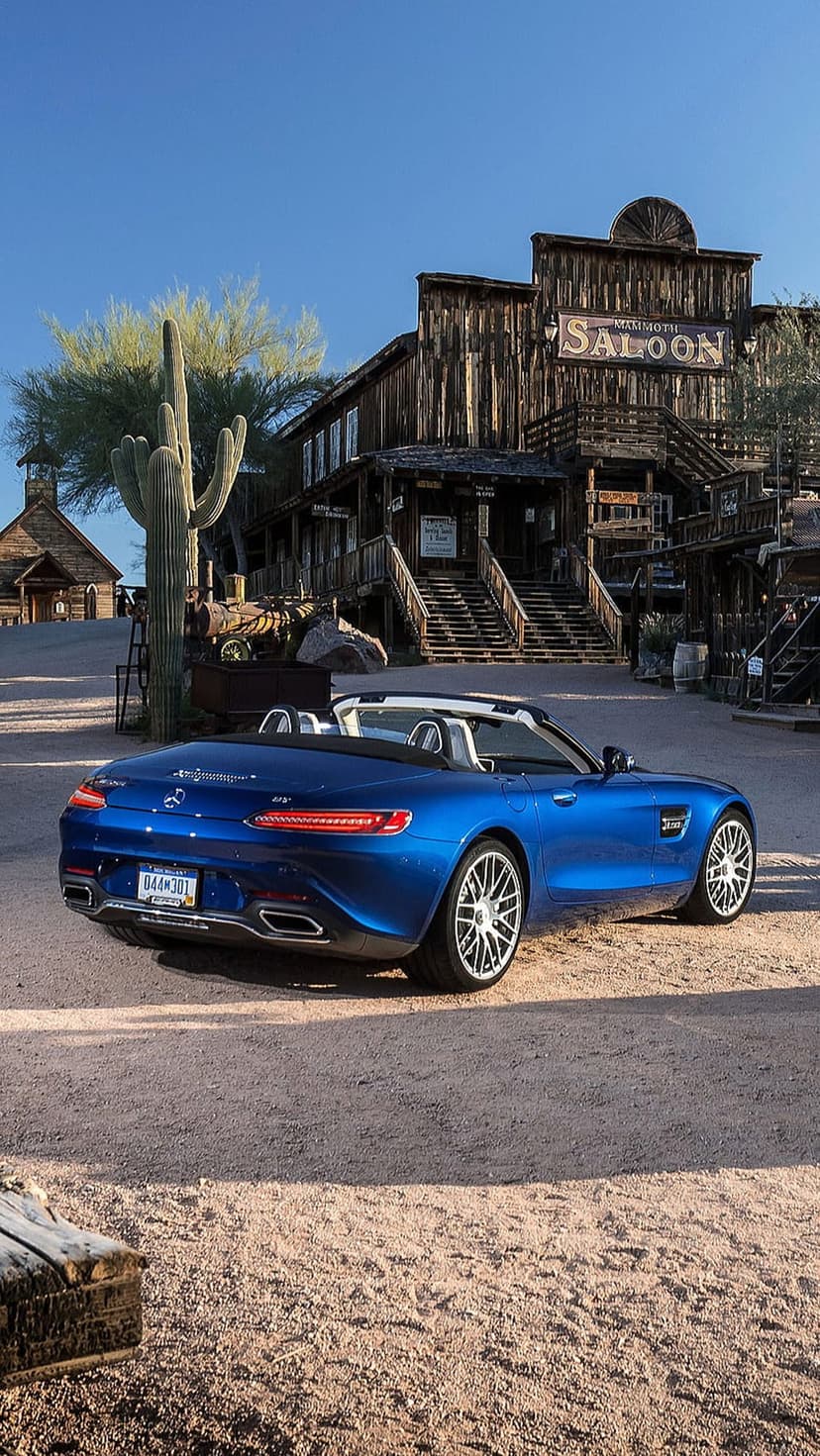 Blue Mercedes convertible parked on a dirt road between rustic wooden saloons and desert cacti under a clear sky.