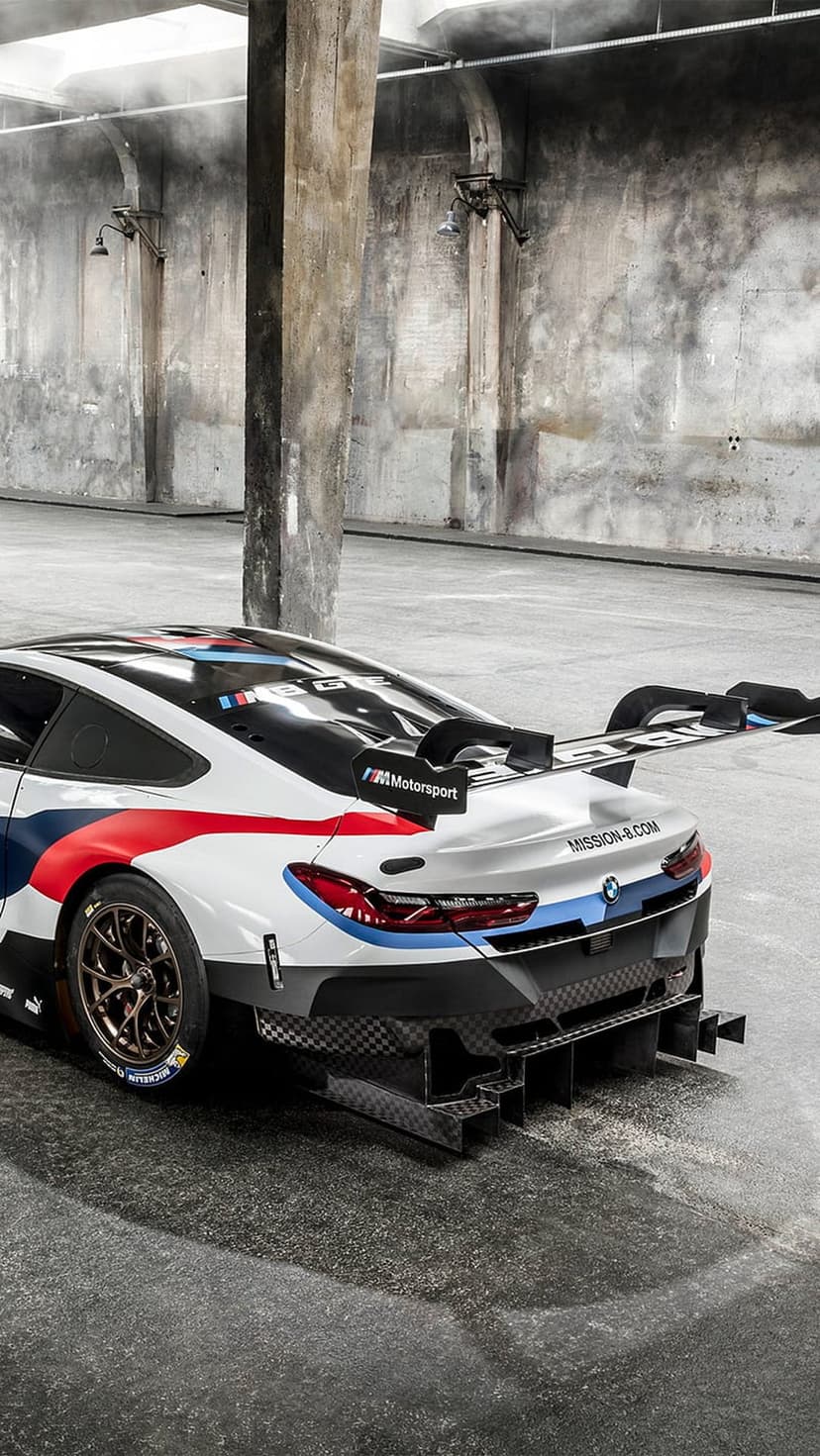 BMW M Motorsport race car with a massive rear wing and red livery parked by concrete pillars in a dark warehouse.