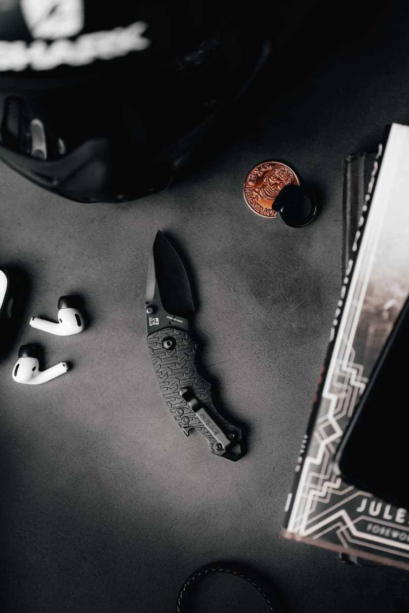 Tactical folding knife, wireless earbuds, and a copper coin rest on a dark book in a moody flat lay composition.