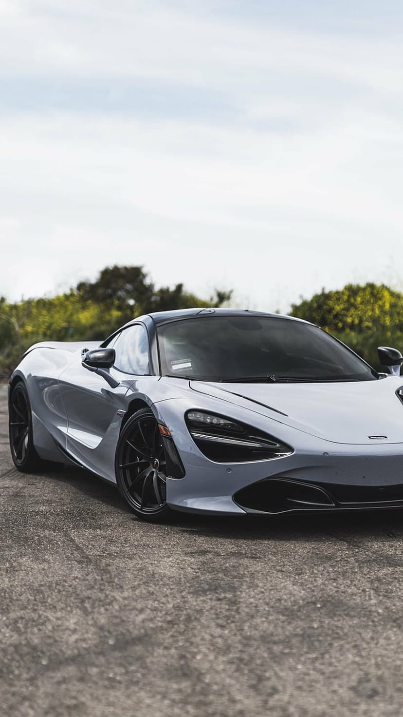 McLaren 720S supercar in metallic grey with black rims parked on asphalt near green trees under a cloudy sky.