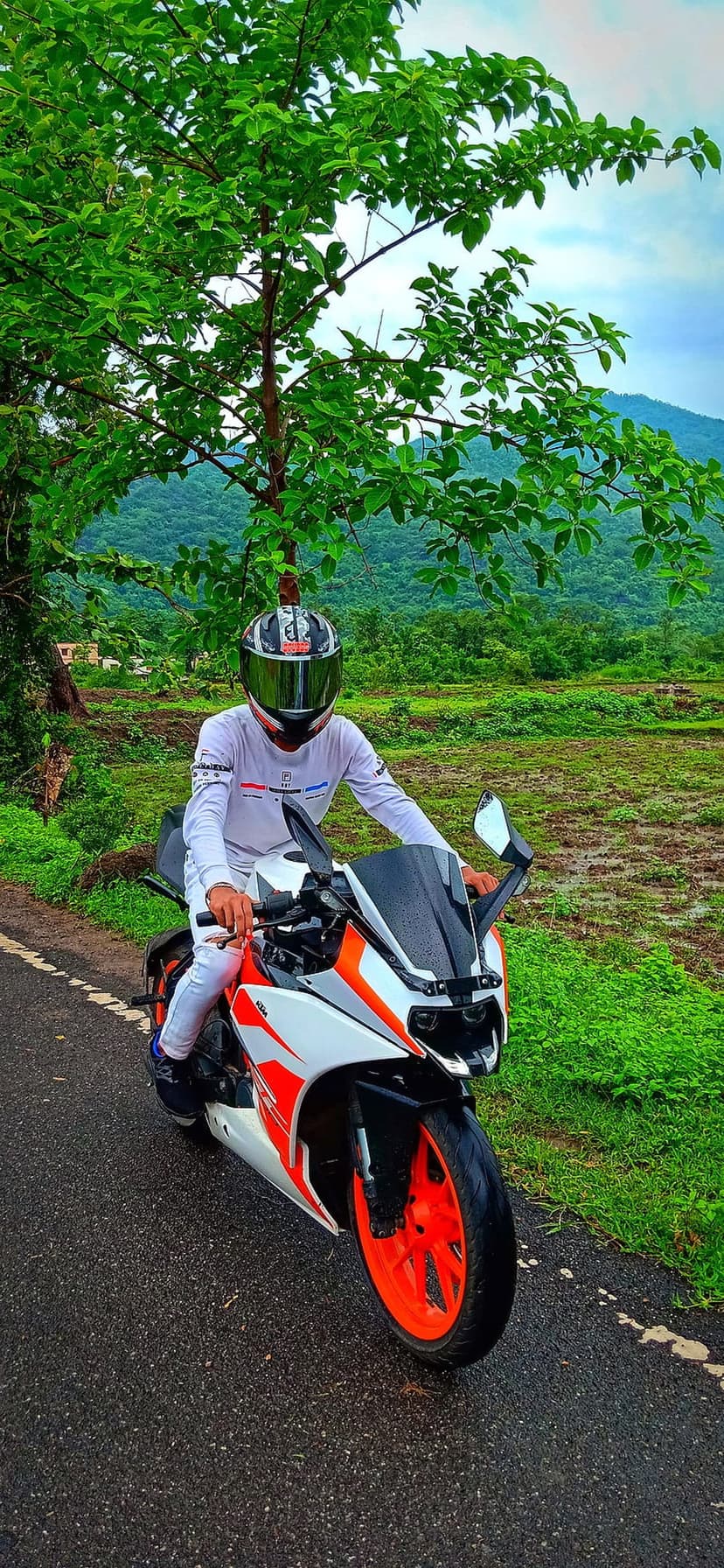 Motorcyclist in a white shirt riding an orange sportbike on a scenic paved road through green forested hills.