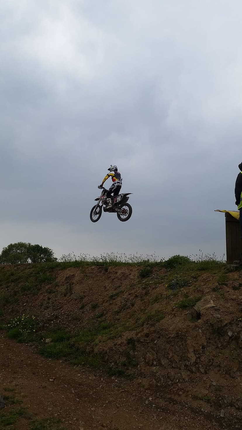 Motocross rider in full protective gear jumps a dirt bike high against a gray and white cloudy sky over a track.