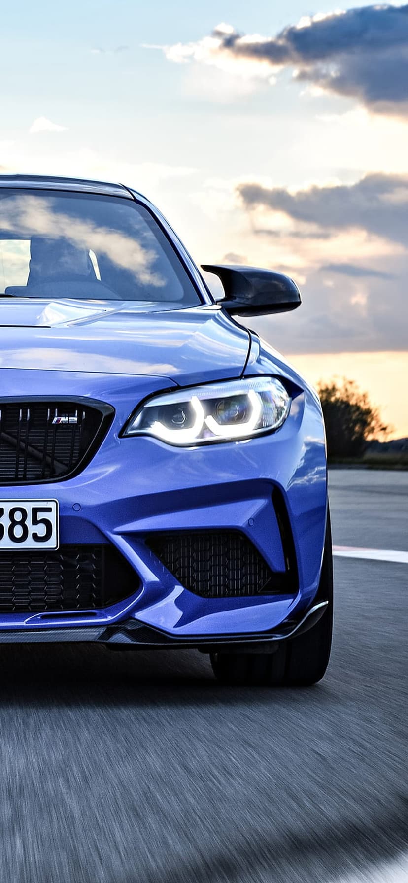 Blue BMW M2 sport coupe driving on a road during a golden hour sunset with motion blur and glowing headlights.
