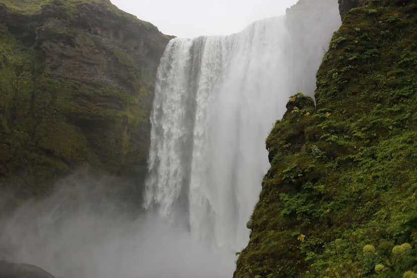 Skogafoss waterfall drops from mossy green cliffs into a misty pool within a vertical Icelandic valley landscape.