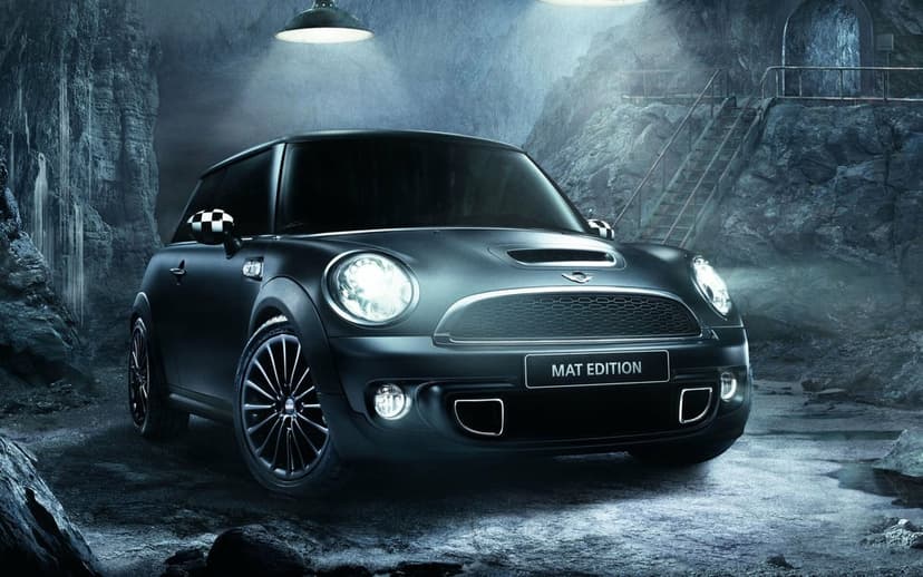 Matte black Mini Cooper Mat Edition with glowing headlights in a dark rocky cave under dramatic spotlights.