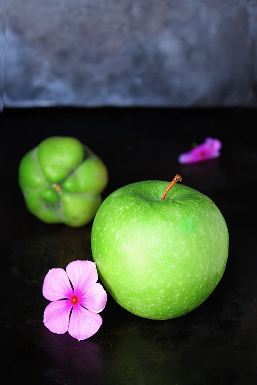 Green apple and a bumpy lime-colored fruit sit on a dark surface next to two small pink flowers.