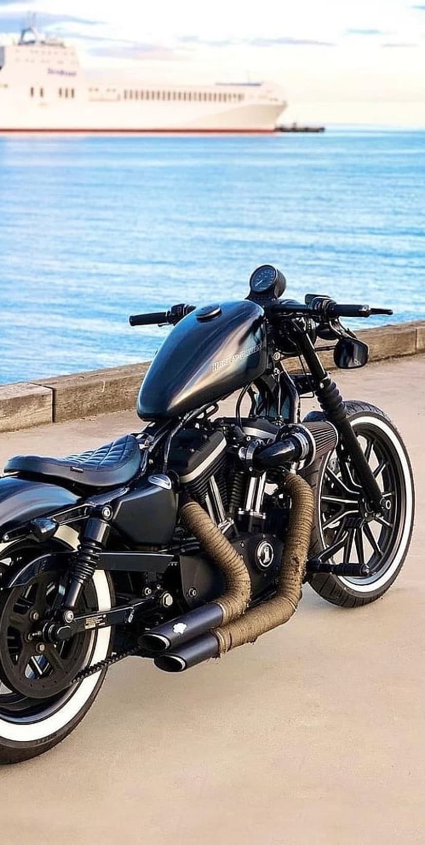 Black Harley Davidson motorcycle with whitewall tires on a promenade with a white ferry boat on blue water.