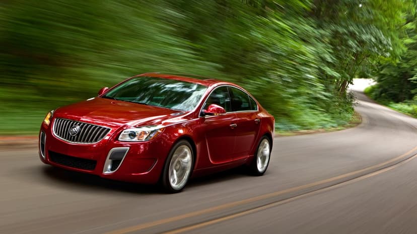 Red luxury sedan driving fast on a winding road through a blurred green forest with spinning silver wheels.
