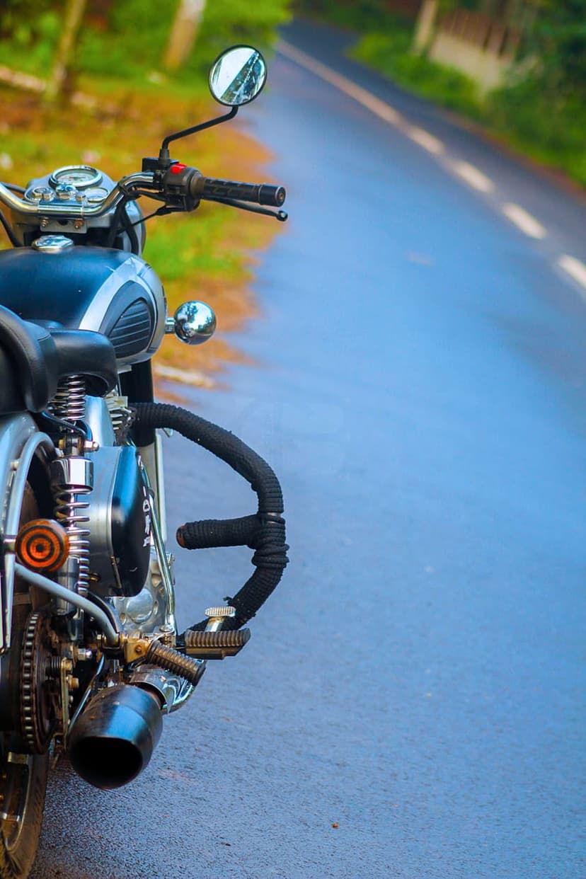 Classic motorcycle handlebars and chrome engine parts focus on a wet asphalt road with a blurred green background.
