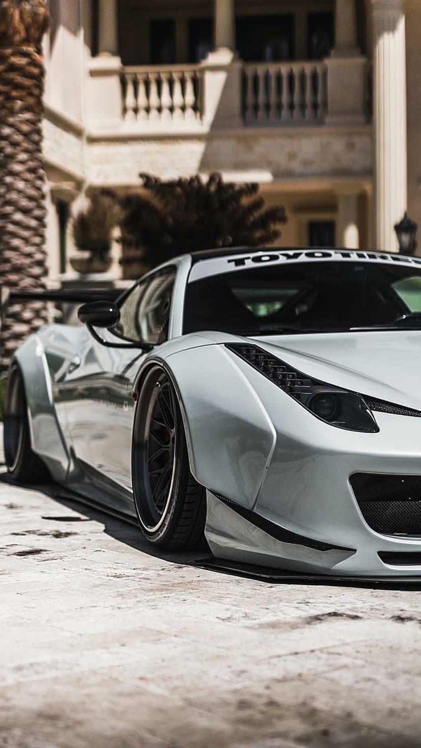 White Ferrari 458 Italia widebody sports car parked on concrete with palm trees and a luxury house behind it.