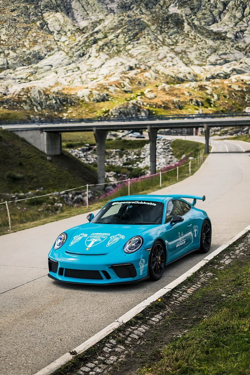 Blue Porsche 911 GT3 with racing decals driving on a mountain road under a concrete bridge in the Swiss Alps.