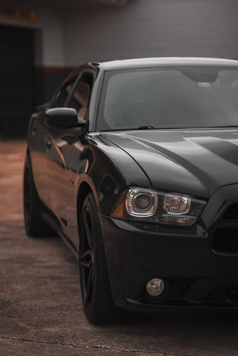 Black Dodge Charger with glowing headlights parked on gray concrete in a moody industrial urban environment.