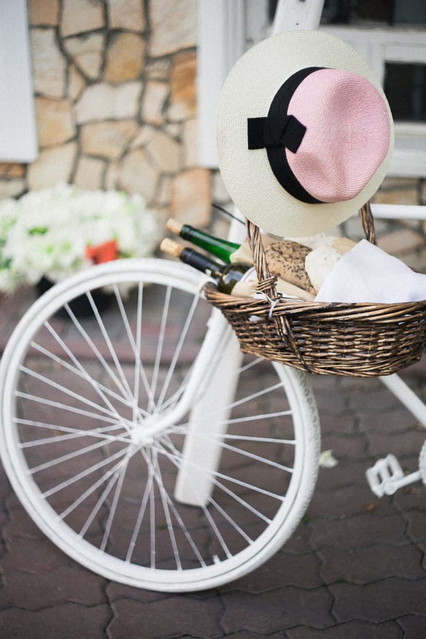 White bicycle with a wicker picnic basket of bread and wine, featuring a straw hat with a pink ribbon crown.