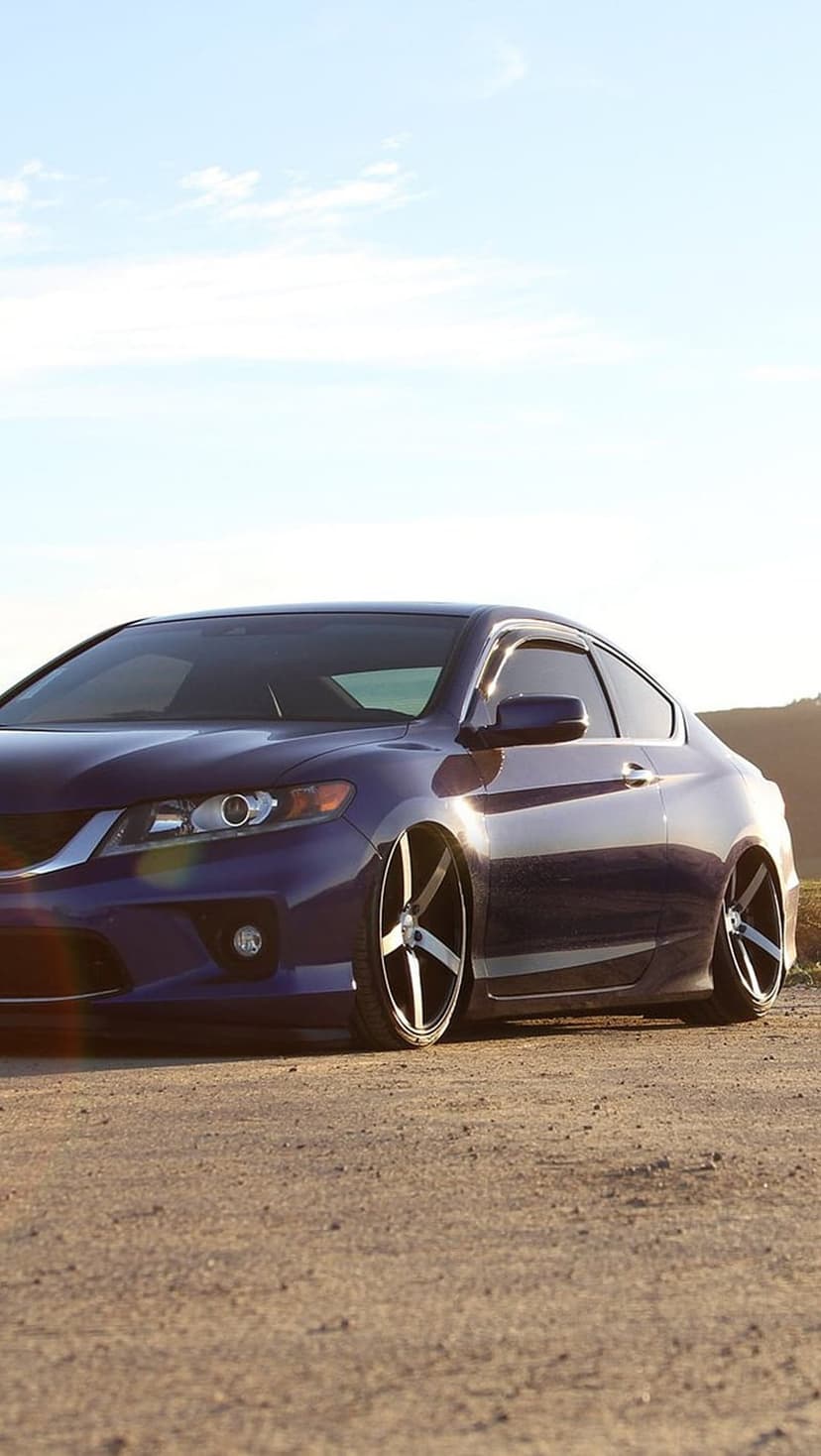 Purple Honda Accord Coupe with aftermarket rims parked on a dirt road with green hills under a bright sky.