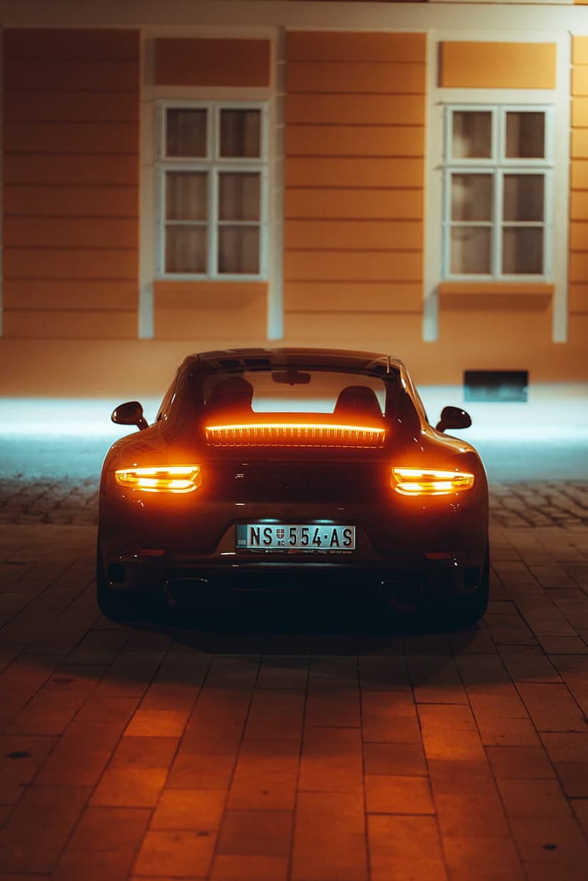 Porsche sports car rear light bar glowing red on a dark cobblestone street with architectural backgrounds.