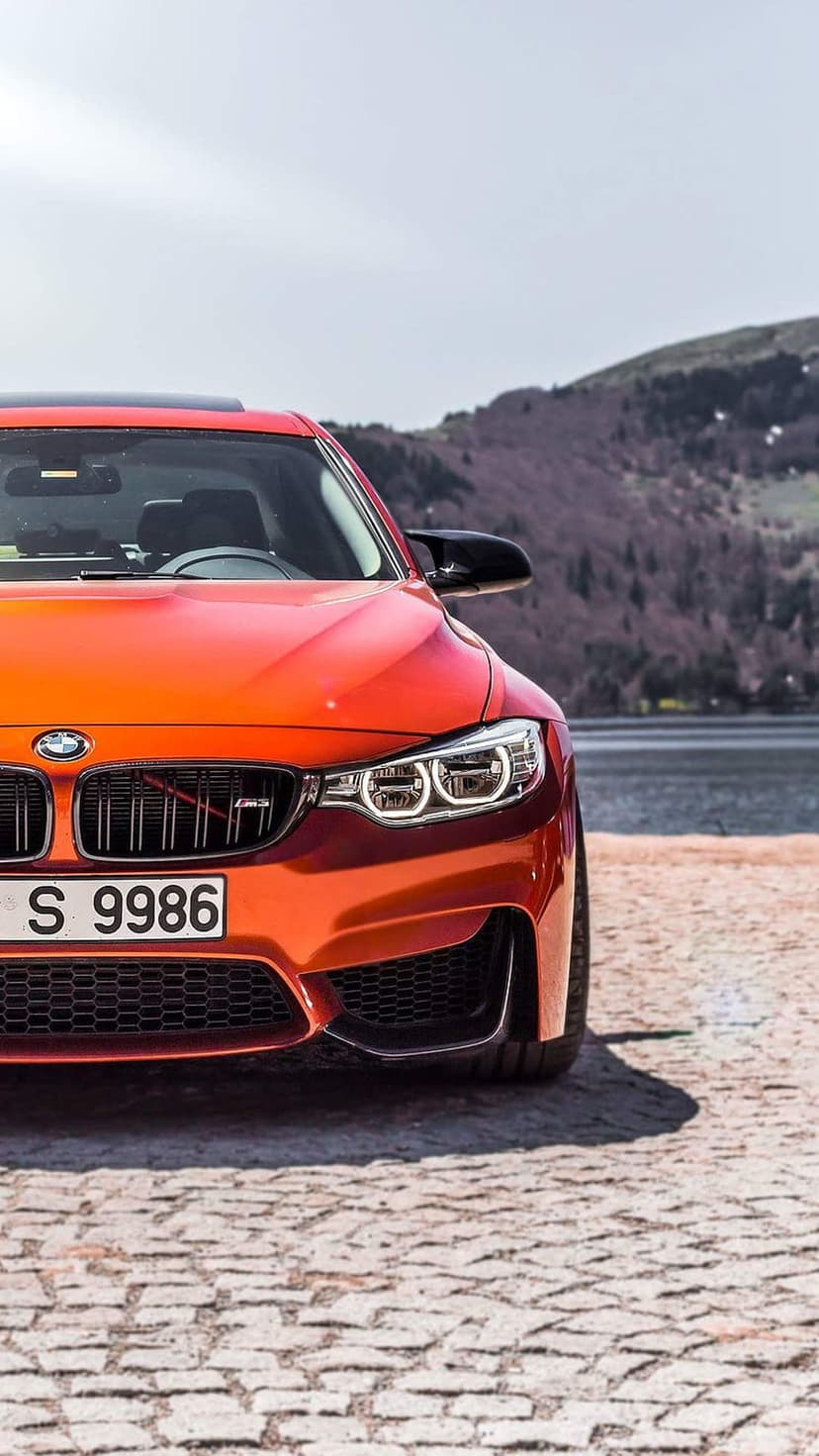 Orange BMW M4 convertible parked on cobblestones with a scenic mountain and lake background in daylight.