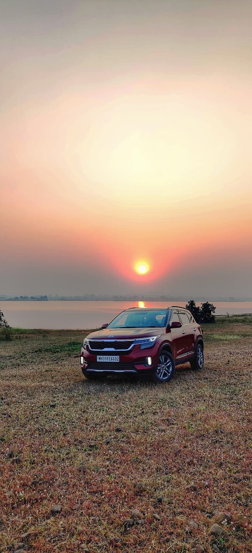 Red SUV parked on dry grass by a calm lake while a vibrant orange sunset reflects on the still water surface.