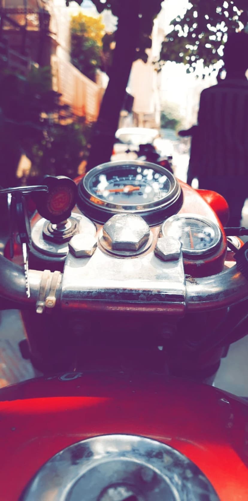Red motorcycle handlebars and a chrome speedometer overlook a blurred urban street with vintage filter tones.
