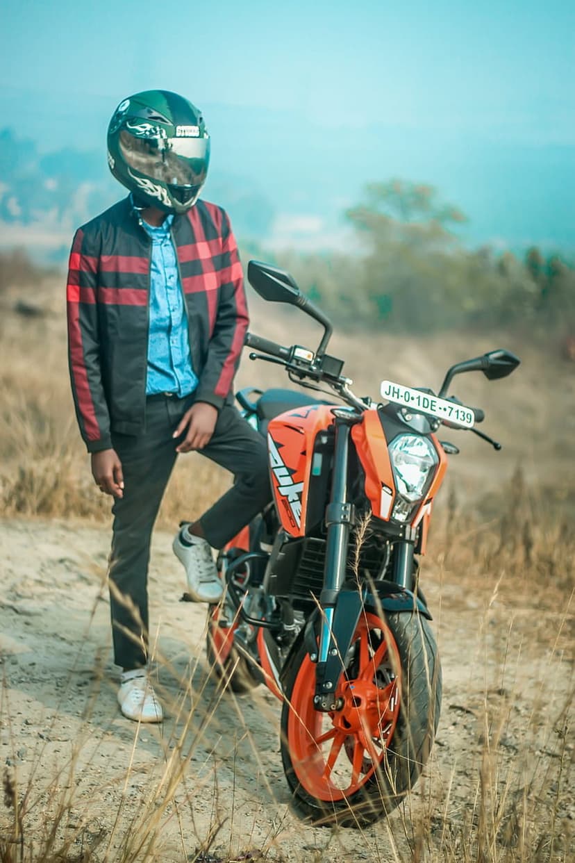 Man in a red checkered jacket and helmet stands next to an orange motorcycle in a field of dry golden grass.