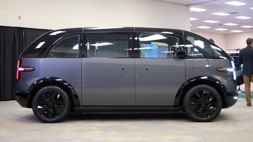 Grey electric minivan with a black top and wheels in a showroom side profile view under bright indoor lights.