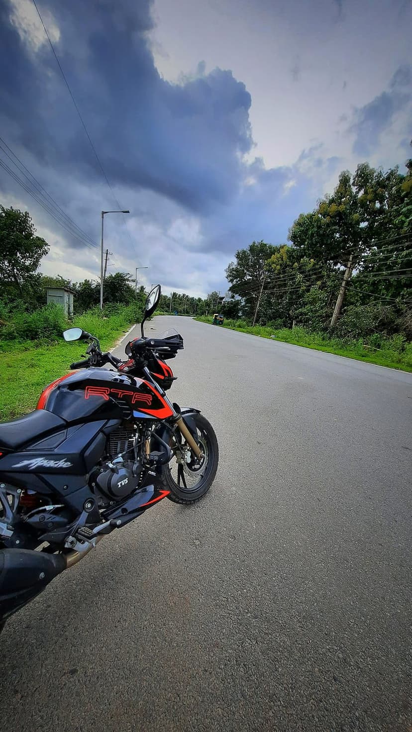 TVS Apache RTR sportbike parked on an asphalt roadside under a dark, dramatic sky with lush green trees behind.