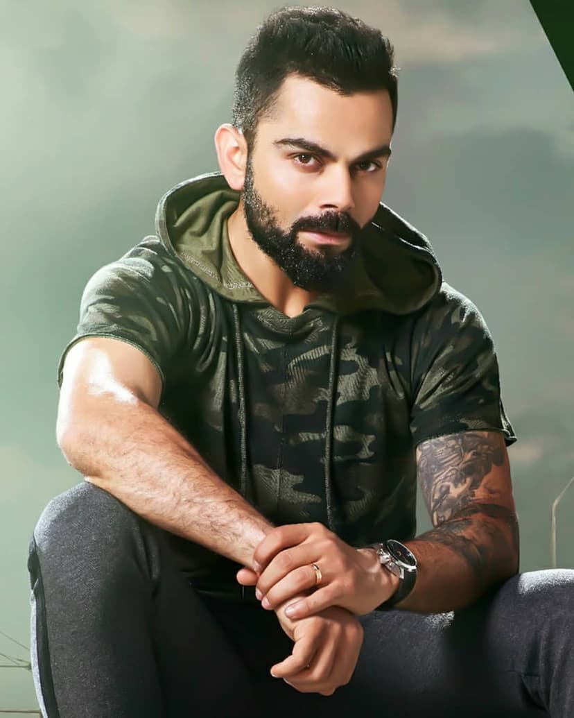 Virat Kohli in a camouflage hoodie and grey track pants with a beard and watch against a soft gray background.