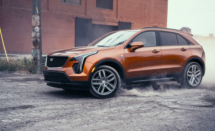 Bronze Cadillac SUV driving on gravel kicking up dust against a red brick industrial building background.
