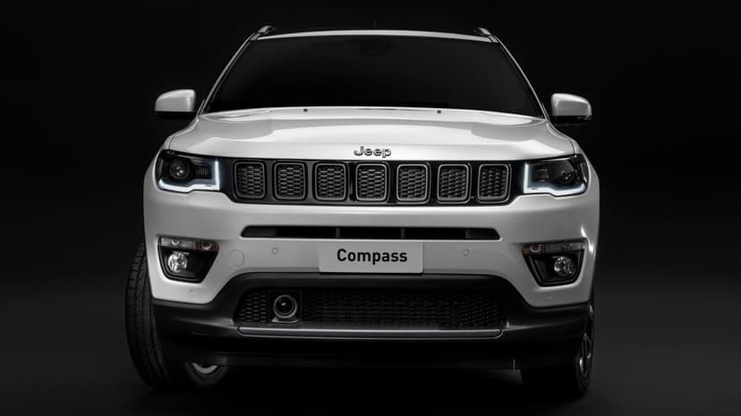 Front view of a white Jeep Compass SUV with illuminated LED headlights against a solid dark studio background.