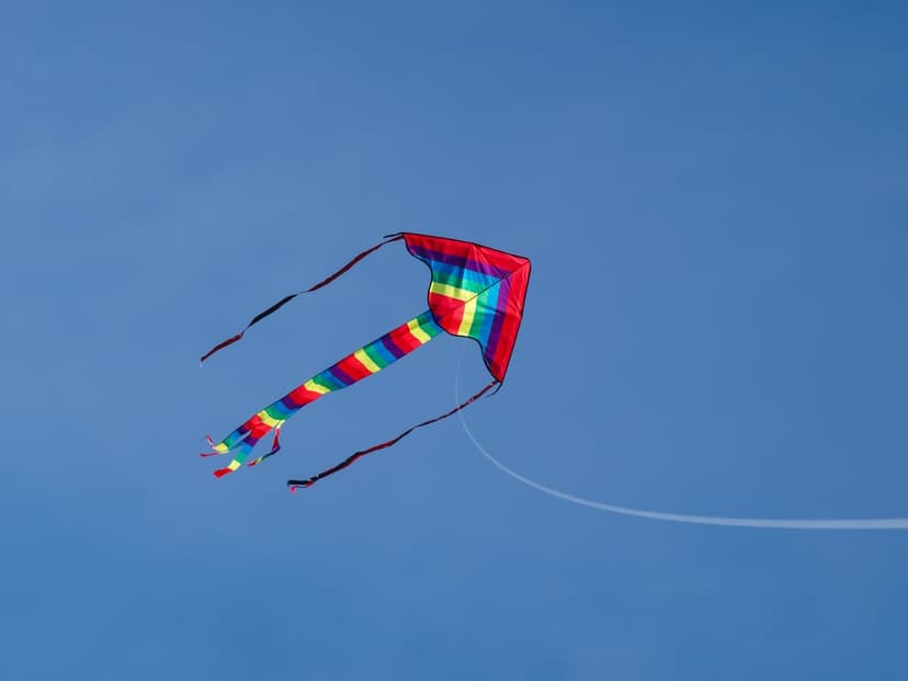Rainbow-striped delta kite with a long flowing tail soaring across a vast and empty bright blue summer sky.
