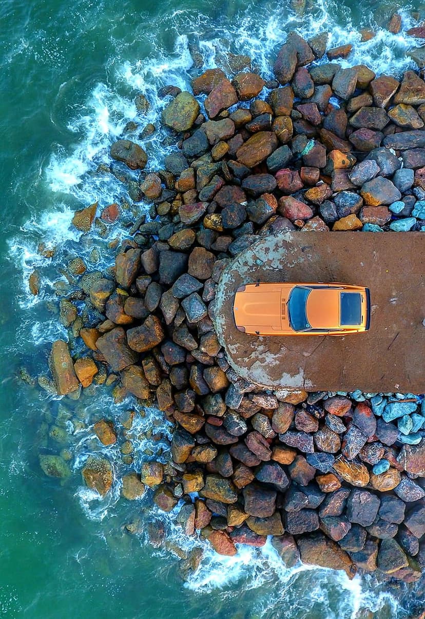 Orange car parked on a concrete ledge above turquoise ocean waves crashing against a dark rocky coastline.