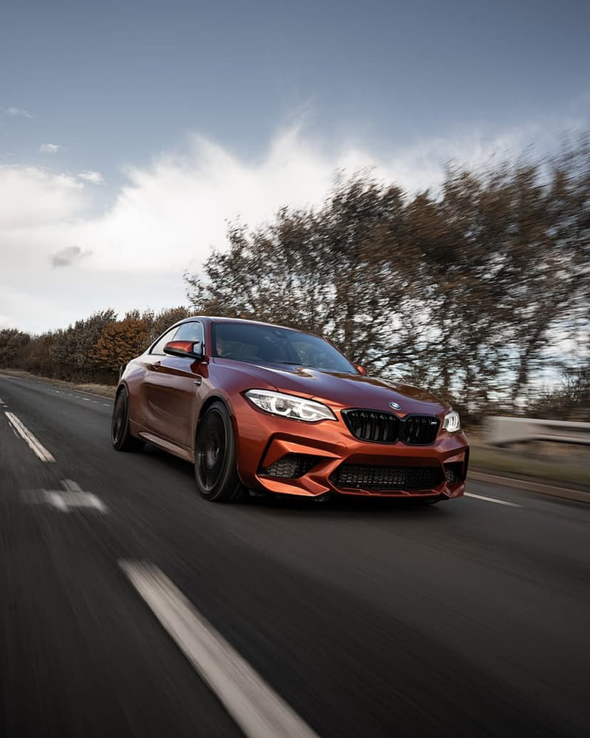 Orange BMW M2 coupe speeds along a highway with motion blur against a background of trees and a cloudy sky.