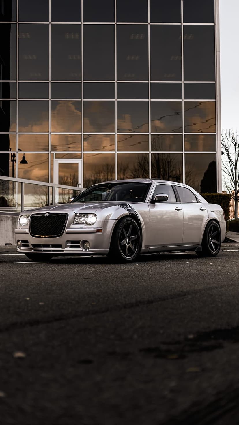 Silver Chrysler 300 sedan with black rims and a fender stripe parked in front of a modern glass building facade.