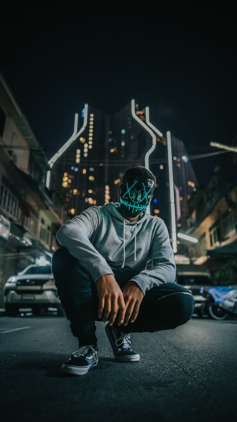 Masked person in a gray hoodie squats on a dark city street with glowing blue X eyes and a jagged mouth.
