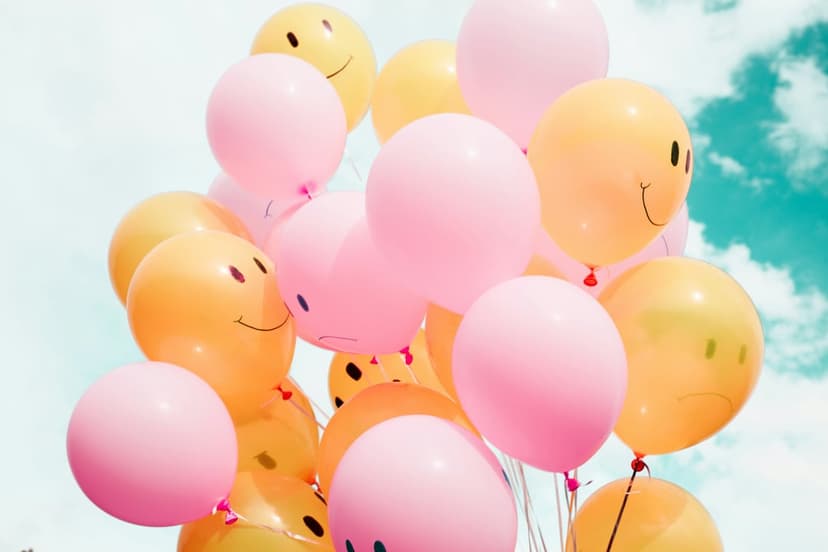 Pink and yellow balloons with smiling and sad faces float against a blue sky with fluffy white clouds in the sun.