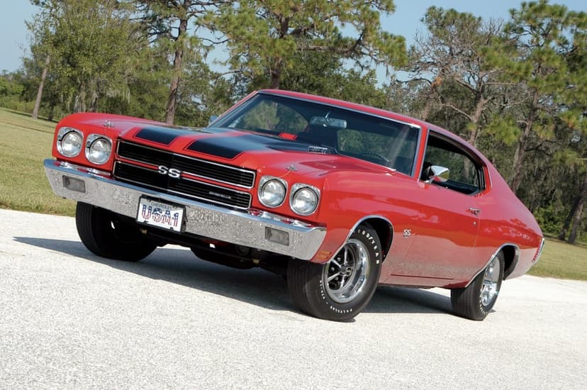 Red 1970 Chevrolet Chevelle SS with black racing stripes parked on a gravel path near green pine trees.