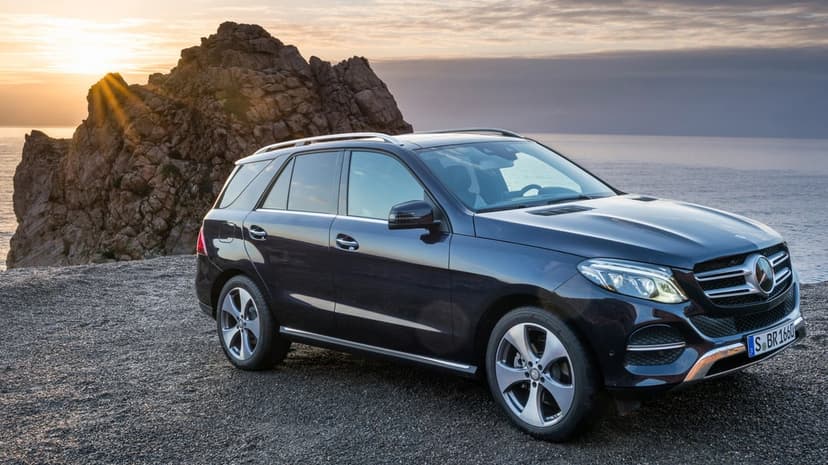 Dark blue Mercedes-Benz SUV on a gravel cliff with a golden sunburst behind jagged rocks by the ocean.