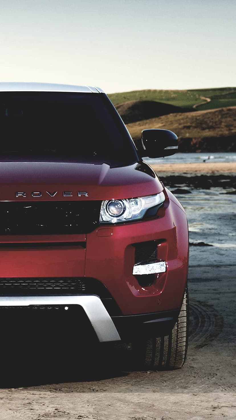 Red Range Rover Evoque SUV parked on a sandy shore with green hills and a bright sky in the background.