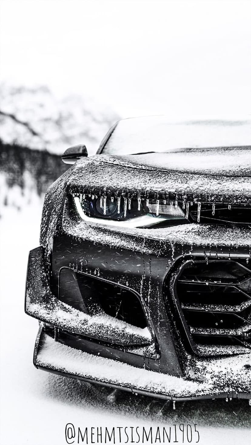 Black car front grille and headlights covered in sharp icicles and thick snow against a blurred winter scene.