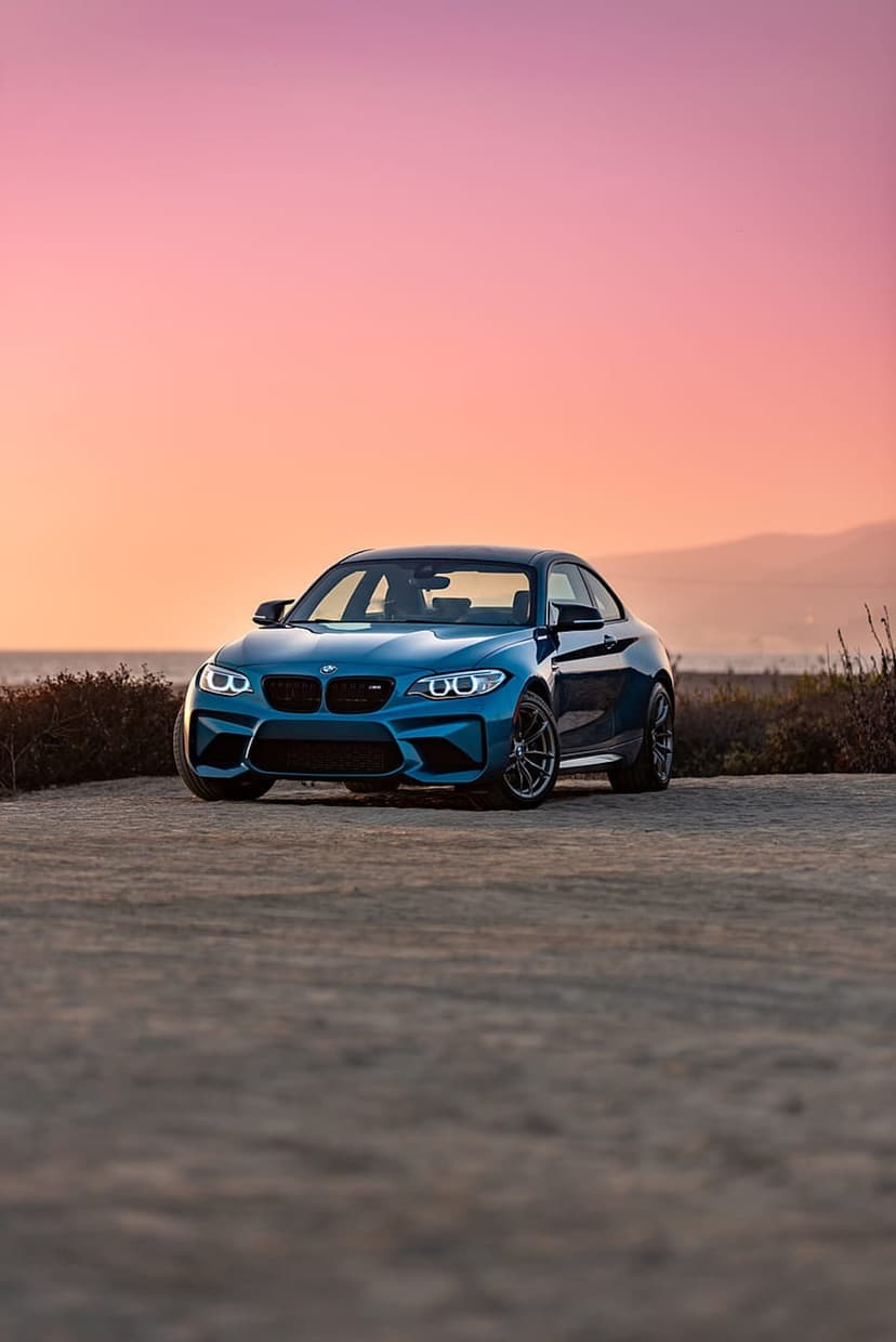 Blue BMW M2 coupe parked on a dirt trail overlooking a calm ocean under a pink and orange twilight sky.