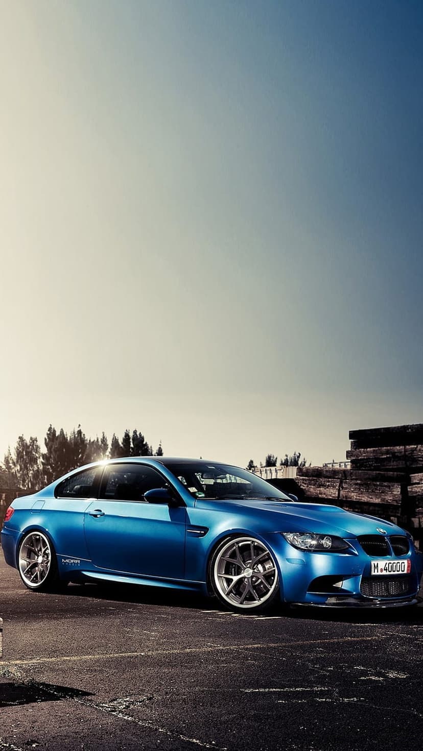 Blue BMW M3 coupe with silver wheels parked on asphalt against a background of trees and a gradient sky.