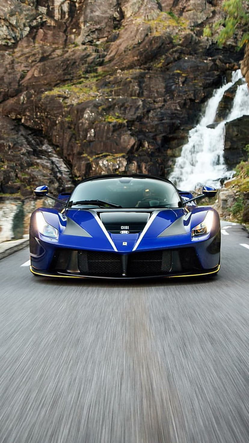 Blue Ferrari supercar accelerates on a wet mountain road next to a cascading waterfall with motion blur effects.