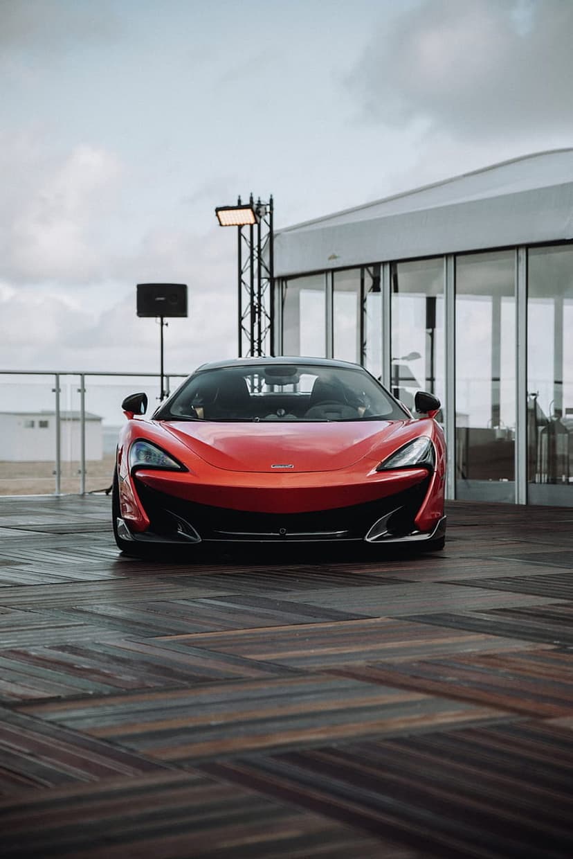 Red McLaren sports car parked on a wooden deck in front of a glass building under a moody, overcast sky.