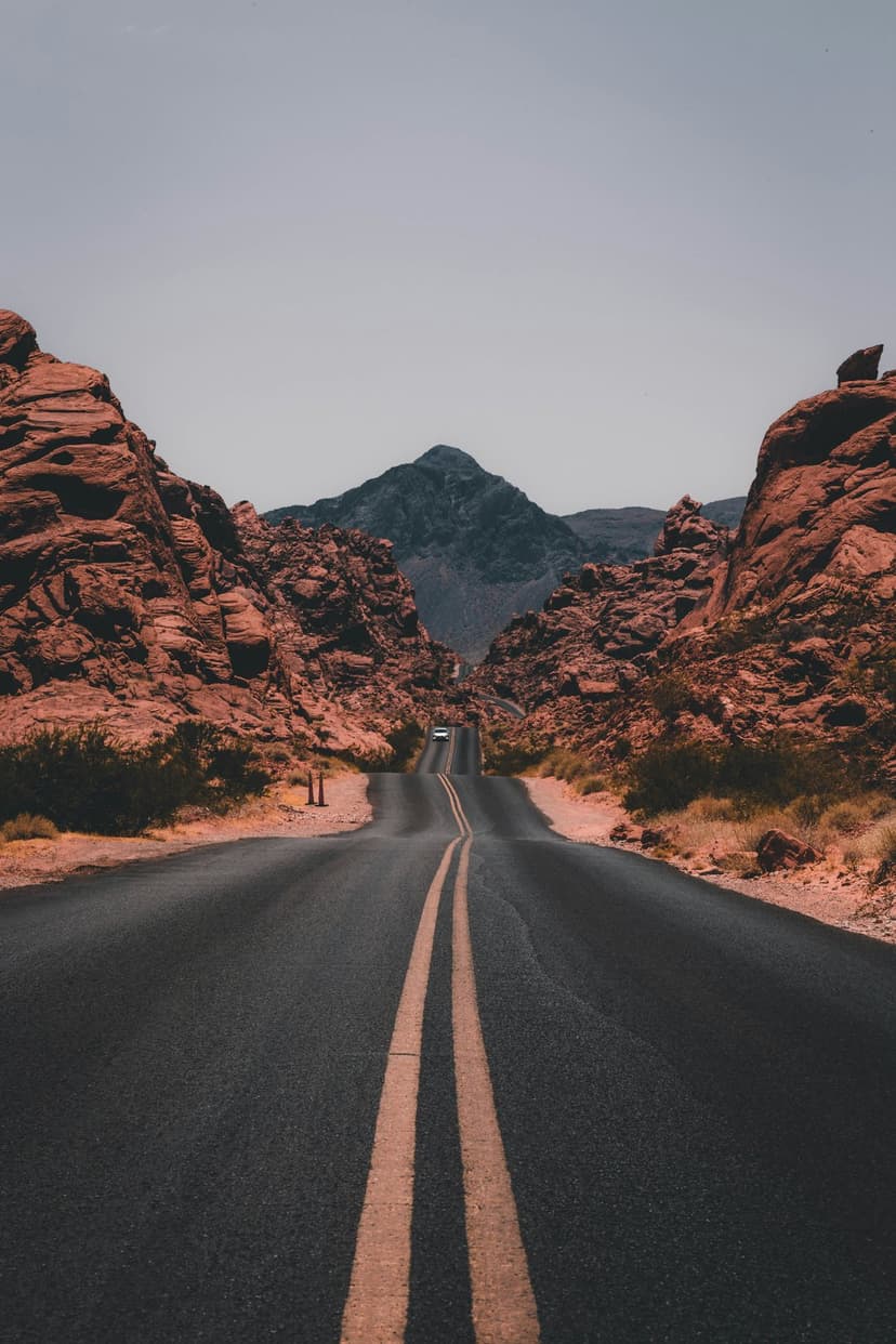 Winding asphalt road with double yellow lines leads past towering red rock formations toward a distant mountain.