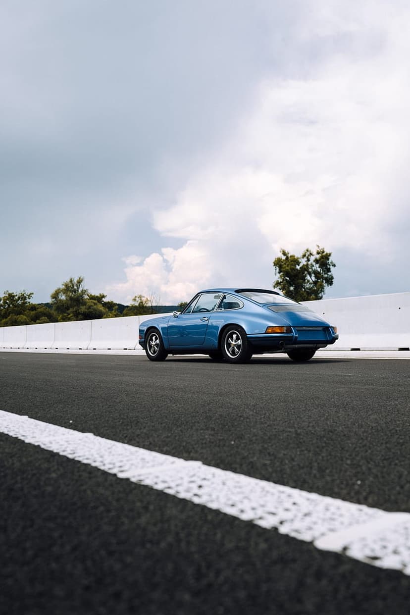 Blue vintage Porsche 911 drives on a wet asphalt road with white markings under a cloudy sky and green trees.