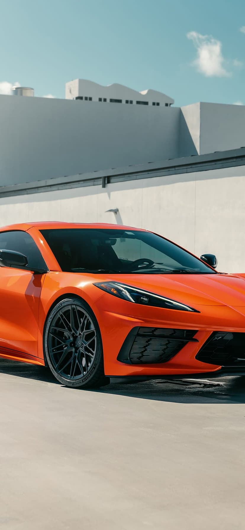 Orange Corvette C8 with black wheels parked before a minimalist white building under a clear blue sky.